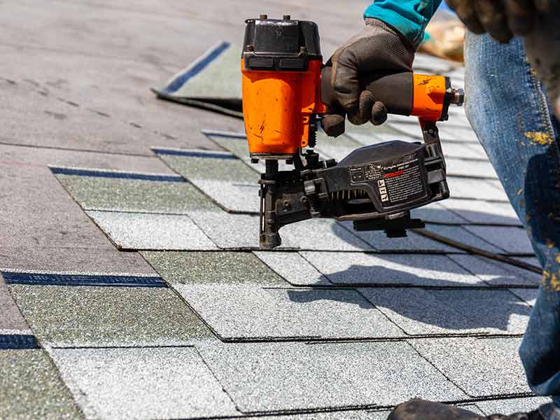 Close up of a worker installing new gray shingles on a residential roof with a nail gun